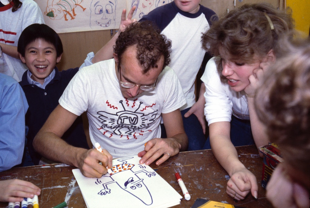 An adult with light skin working on a drawing surrounded by kids and adults.
