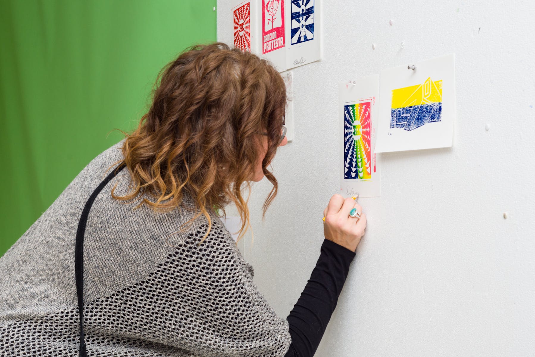 An adult with long hair and light skin writes on the wall next to a colorful print with a pencil.