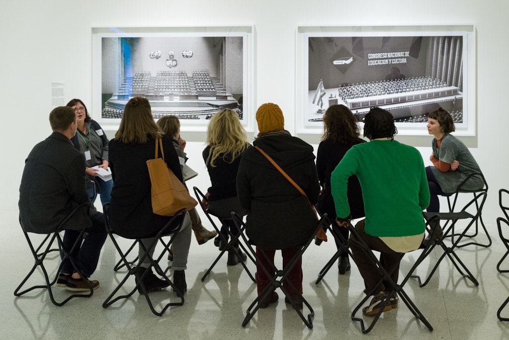 A group of adults sit in chairs in front of photographs hung on a gallery wall.