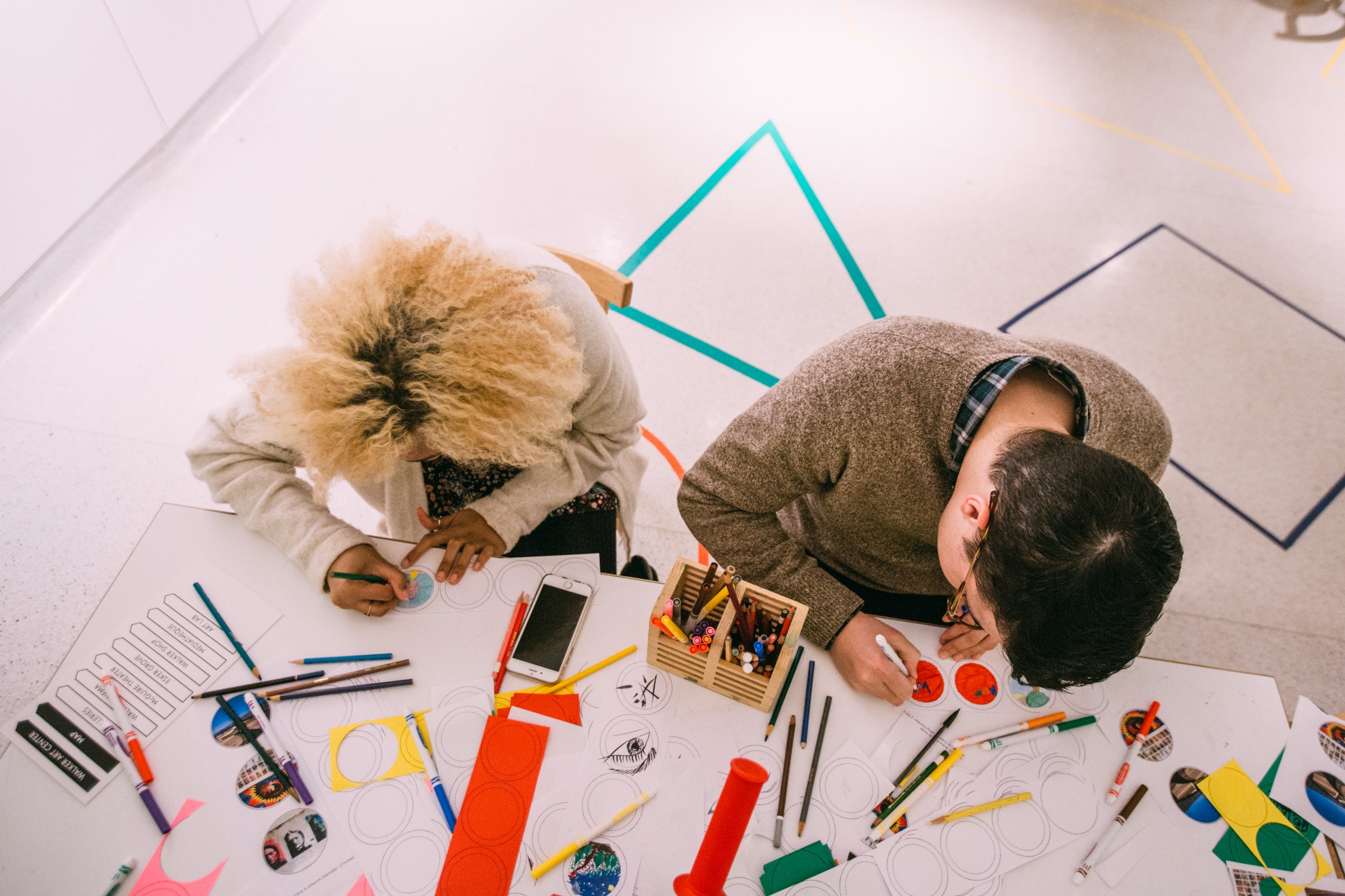 Two people sit at a table drawing and coloring with markers and craft supplies scattered around.