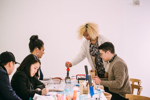 A group of five young adults sit at a table full of crafts one adult is standing and has curly blonde hair using a tool on the table.