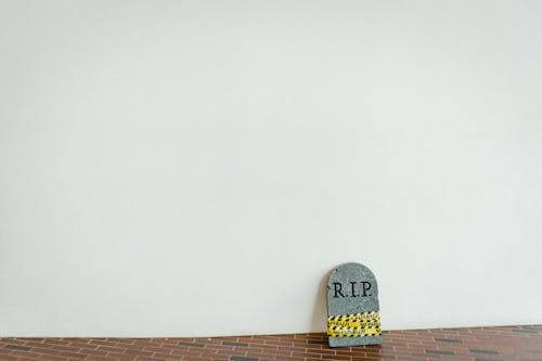 A white wall and a small faux gravestone on the floor that says "R.I.P" and is wrapped in caution tape.