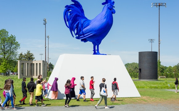 A tour group walks past a large blue rooster sculpture outside on a sunny day.