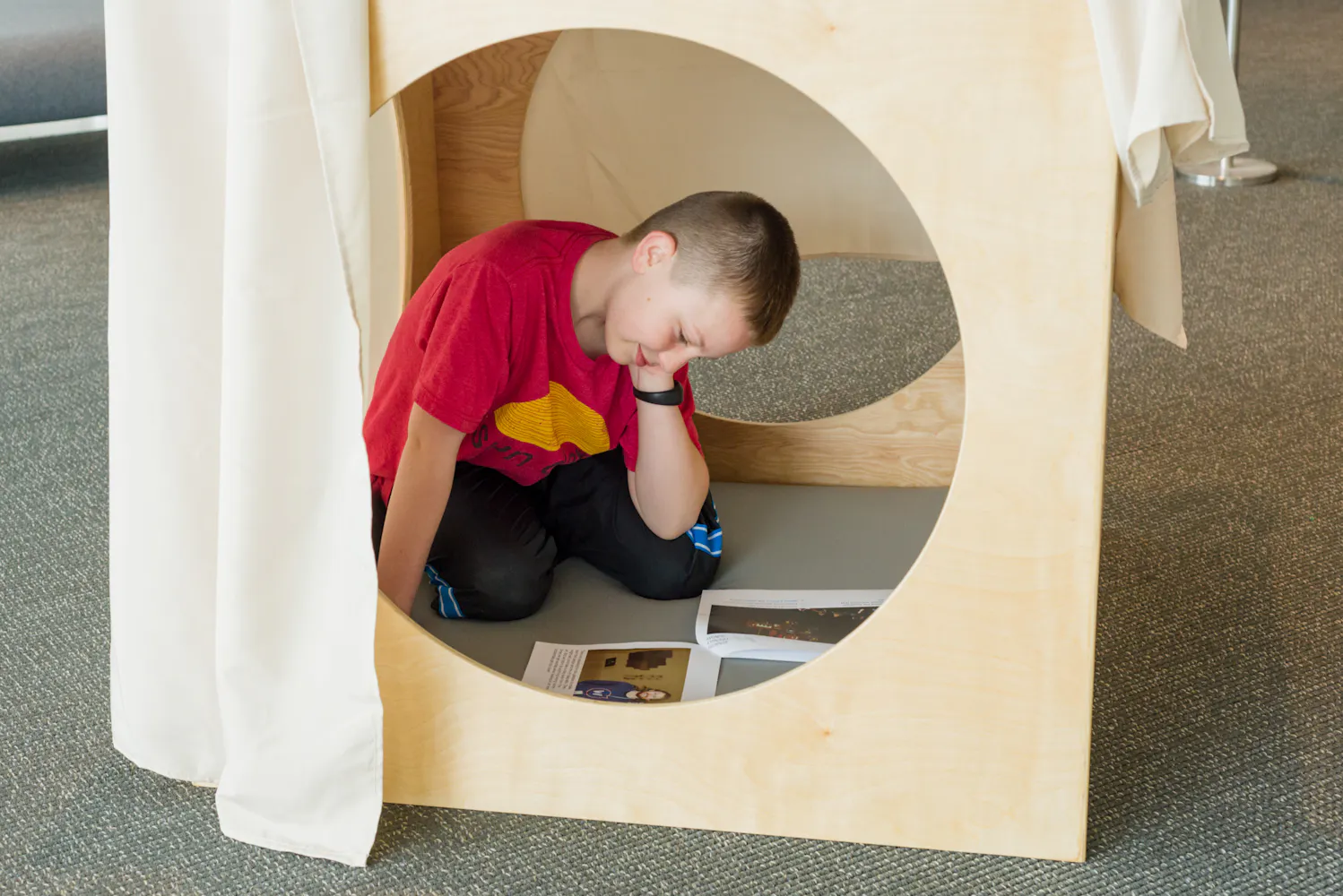 A boy sides inside a wooden box with circles cut out and looks at a patcket of pictures.