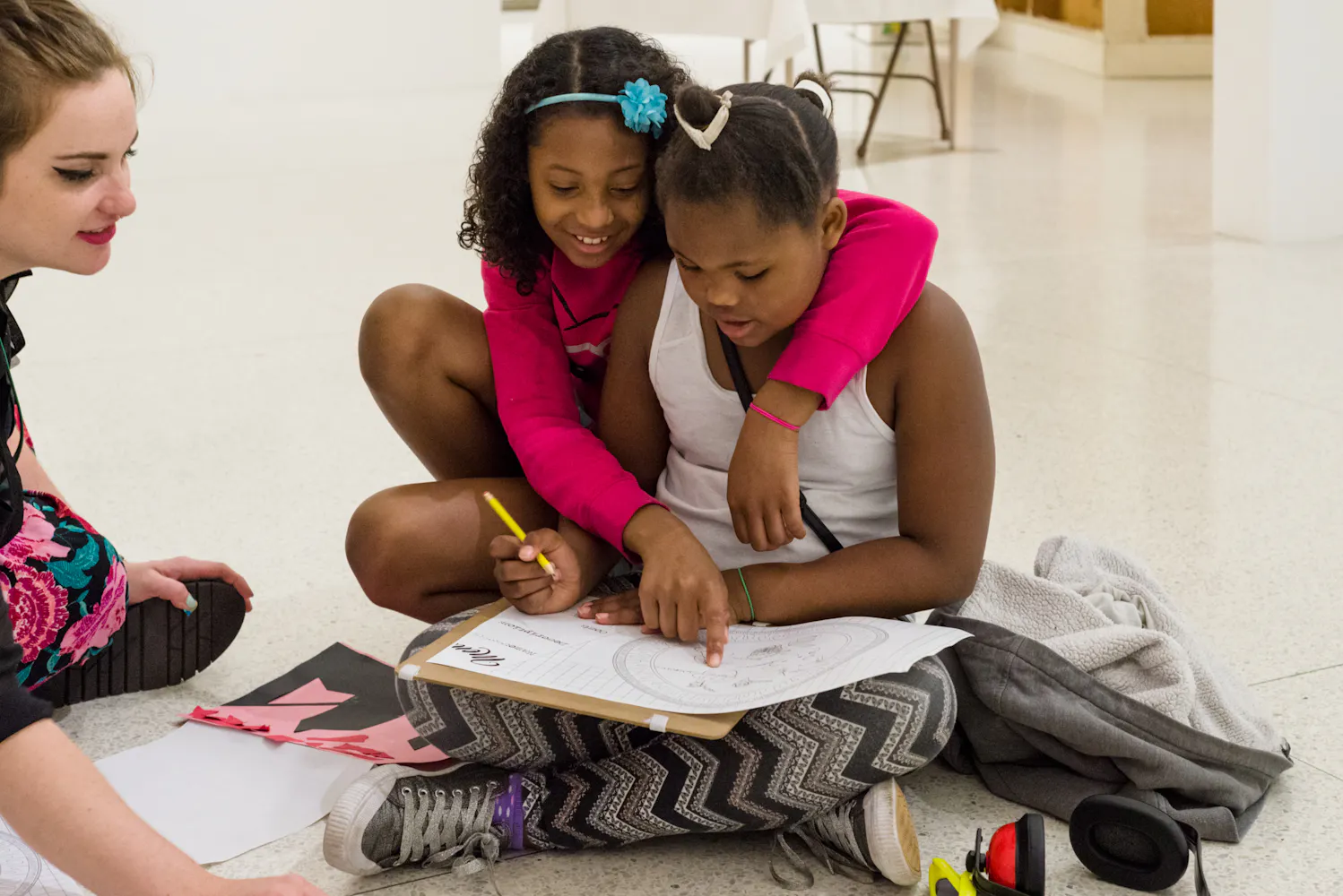 A girl sits on the floor drawning while another girl sits next to her with her arm around her.