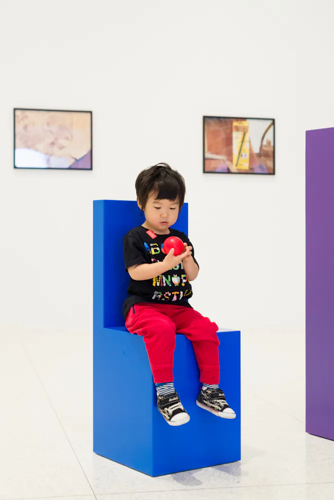 A young boy sits on a custom blue, blocky chair while holding a red ball in his hands.