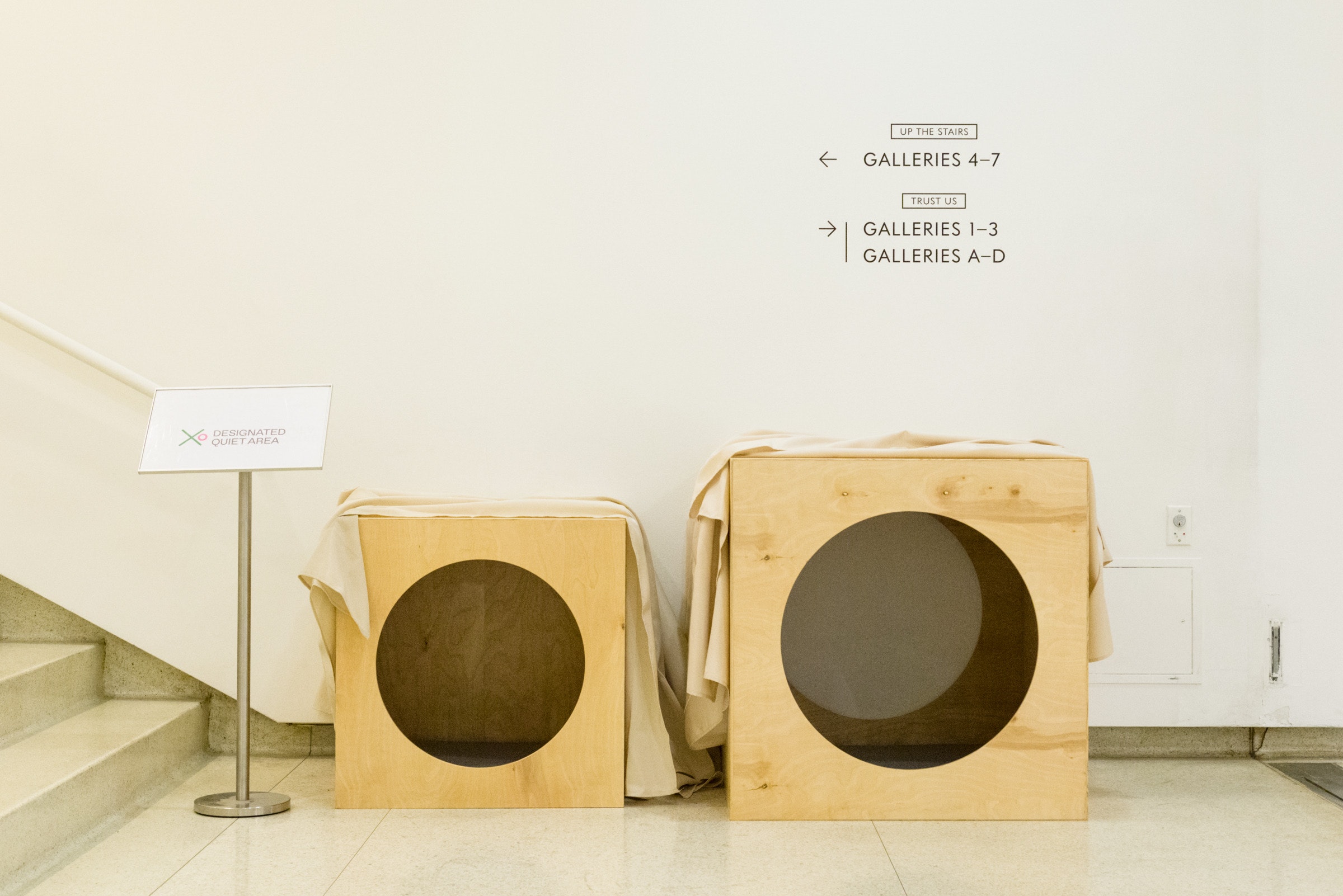 Two woodend boxes with circles cut out on all sides sit in a gallery next to sign for their use as play areas.
