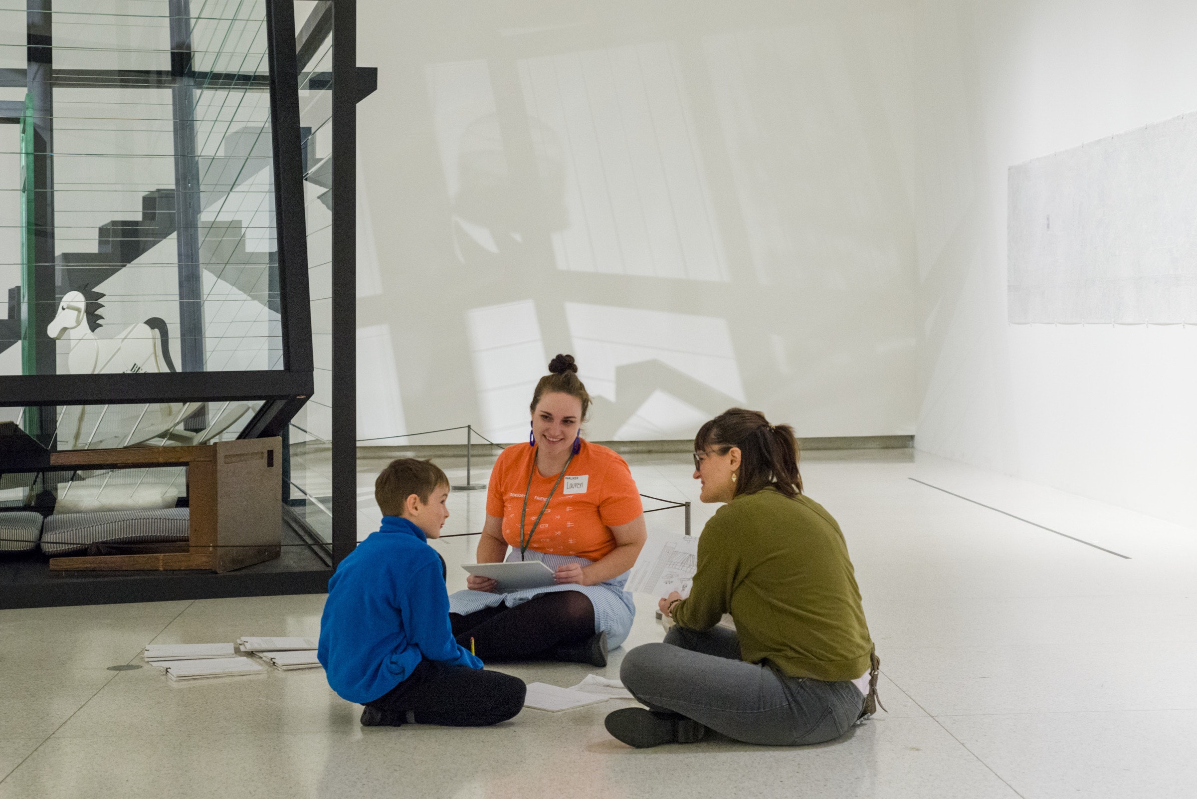 Two women and a boy sit on the floor of a gallery talking to one another.