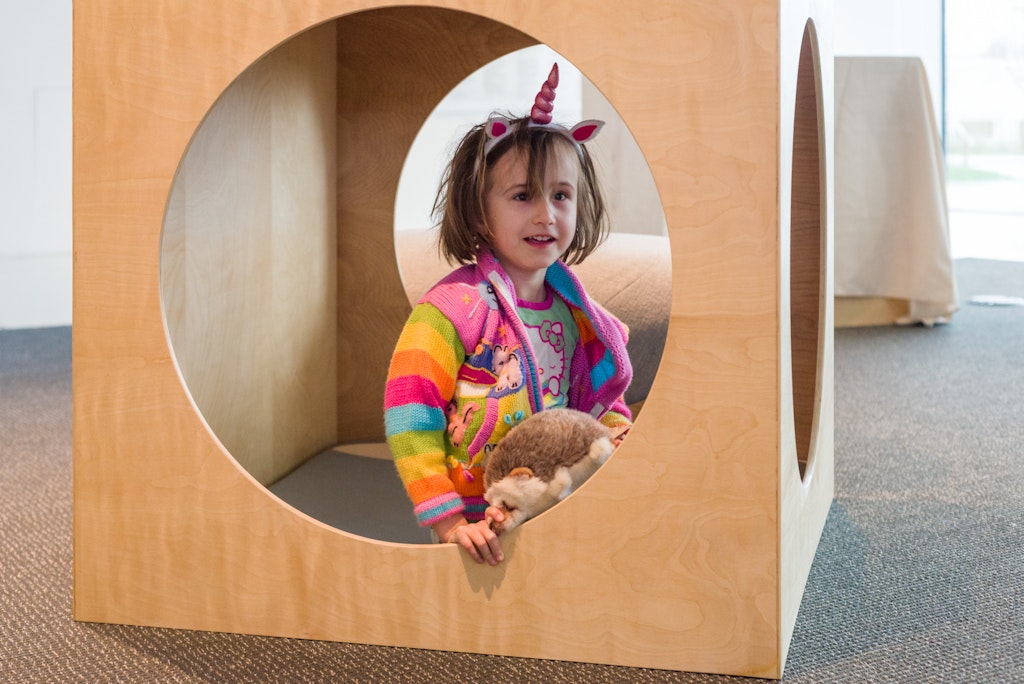 A young girl wearing stuffed unicorn horn and ears plays in a wooden box with circles cut out on all sides.
