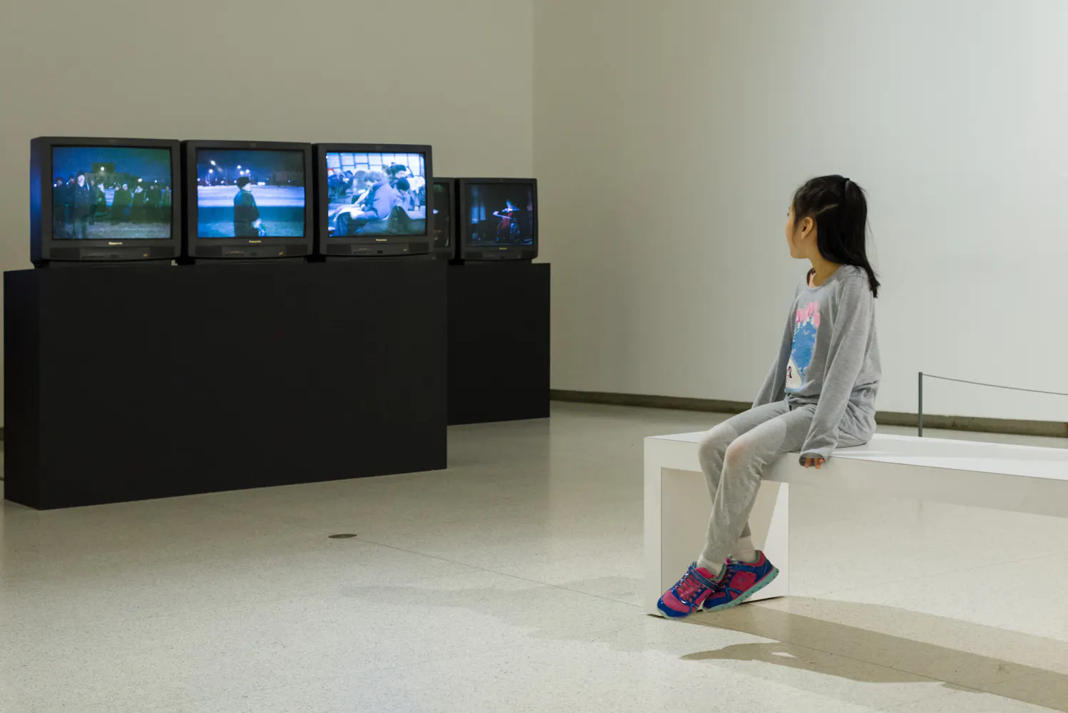 A young girl sits on a gallery bunch and watches several TVs.