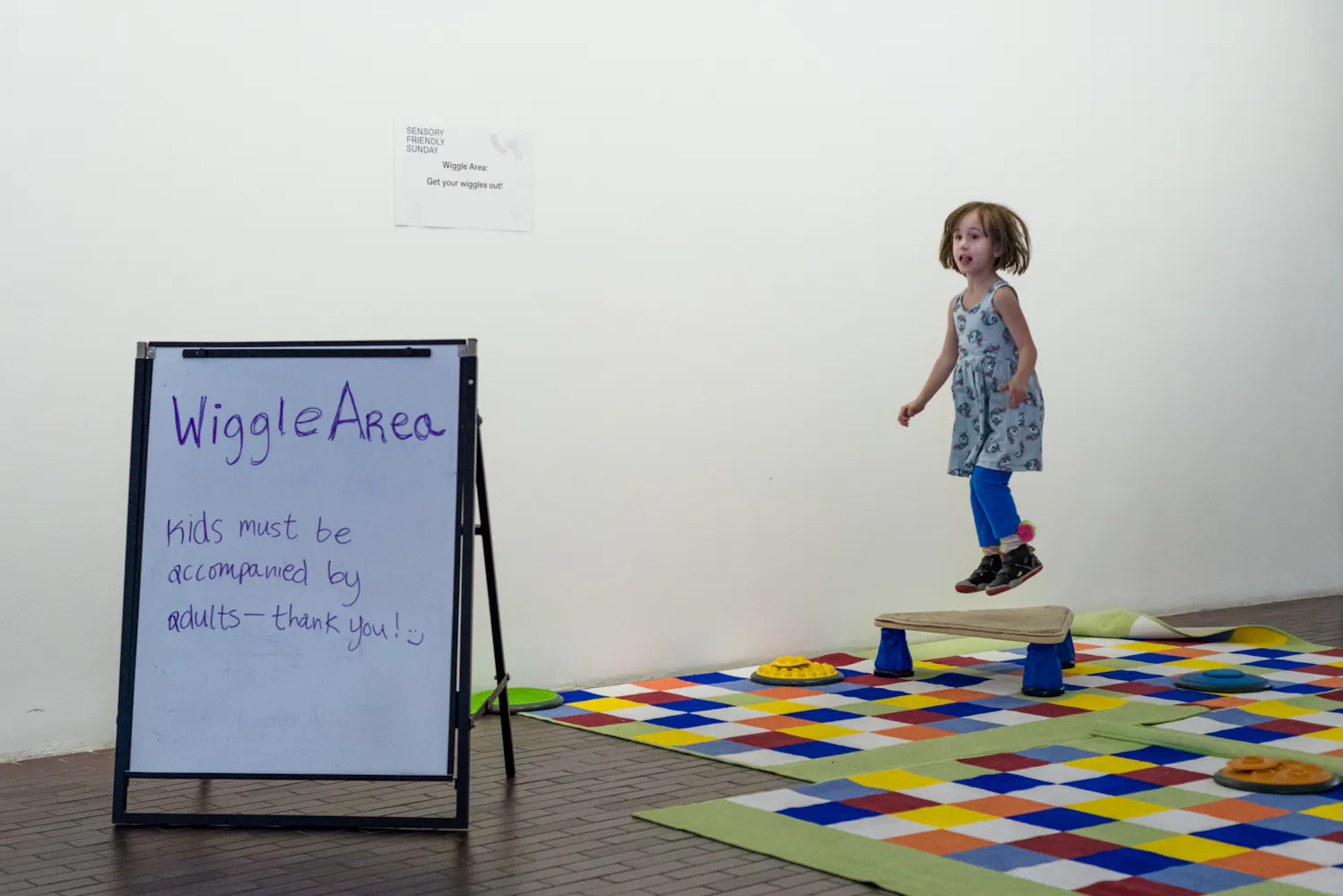 A young girl jumps in the air in a section of a museum desiinagted a wiggle area by a nearby sign.
