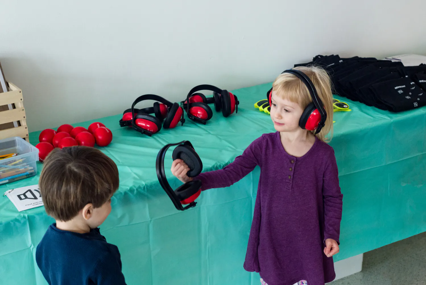 A young girl wearing noise cancelling headphones hands another pair of headphones to a young boy next to a table with many headphones.