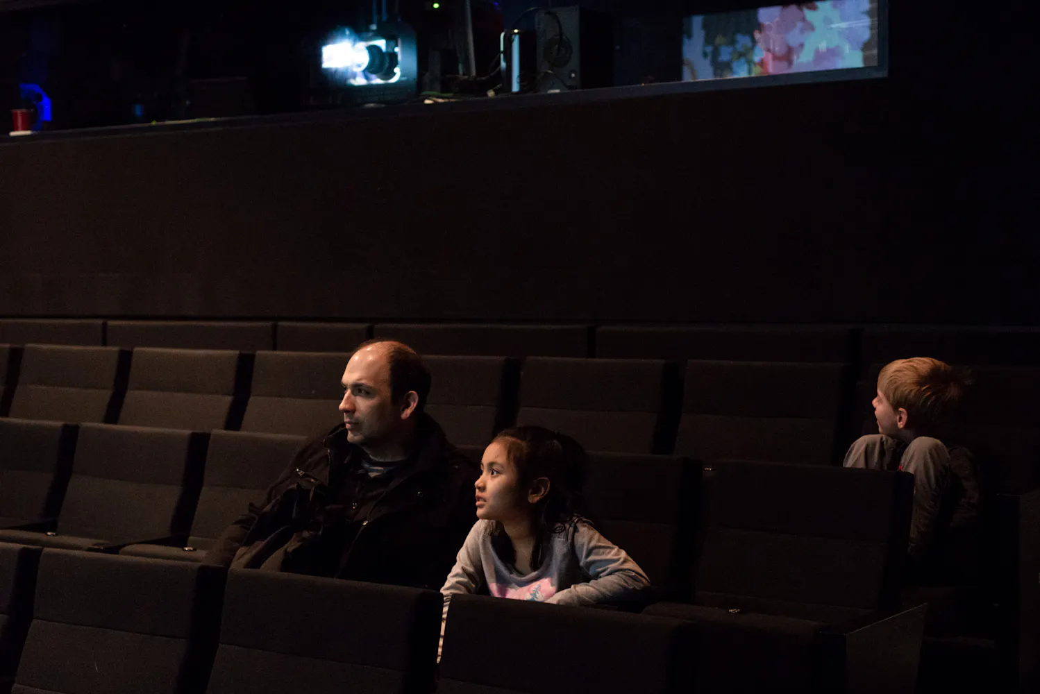 A man and two children sit in a darkended theater watching a movie.
