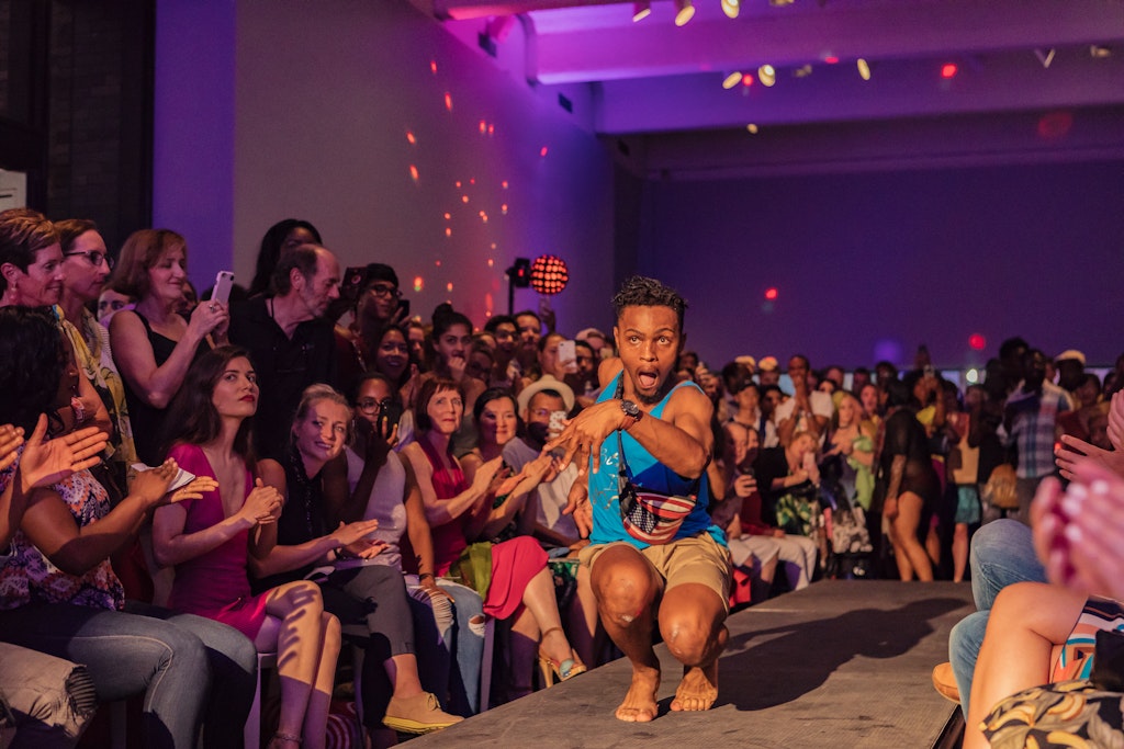 An adult vogue dances down a catwalk surrounded by a cheering audience inside of the Walker Art Center.