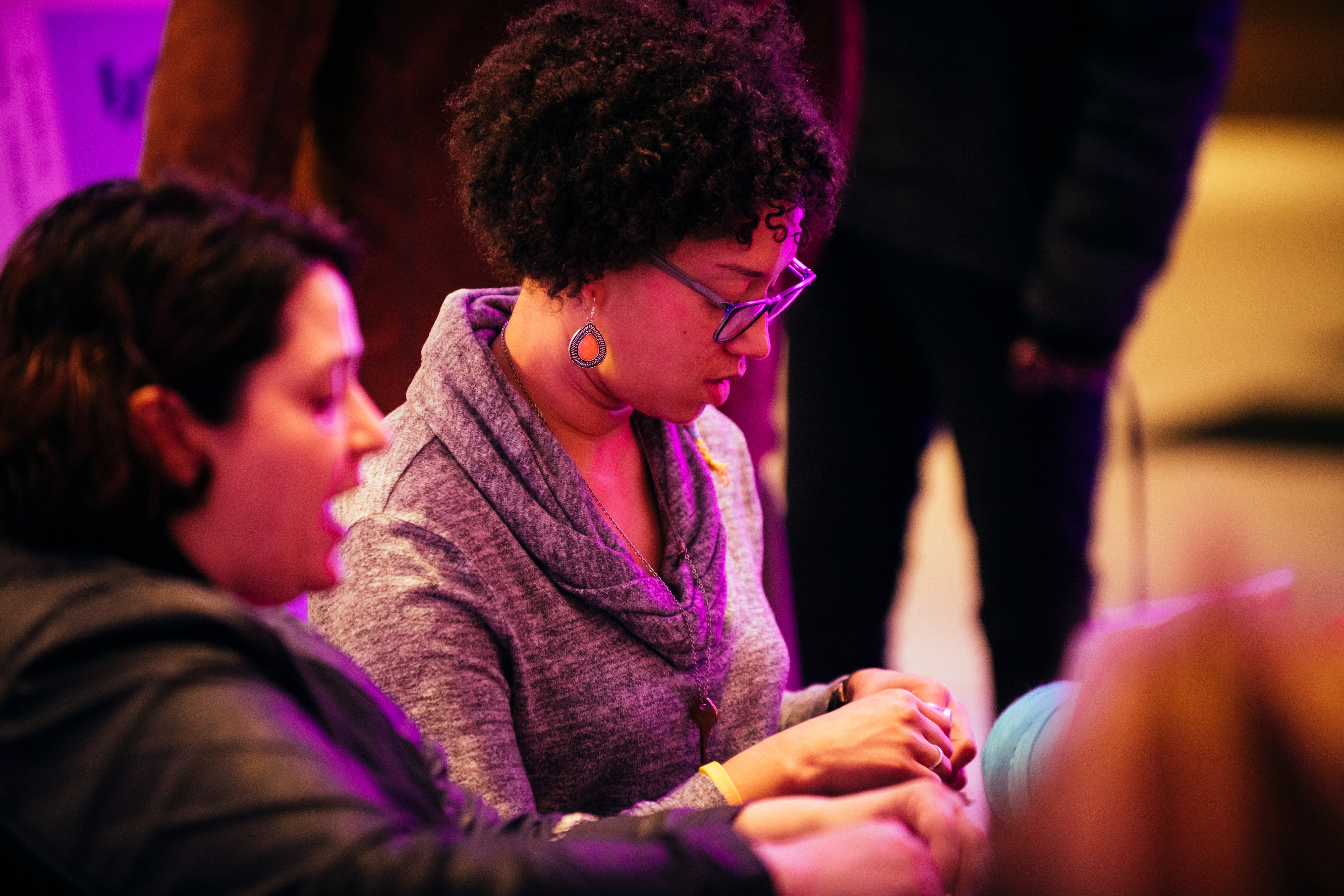 An adult with curly black hair and classes, looks down at something in their hands, a out of focus person sits next to them.