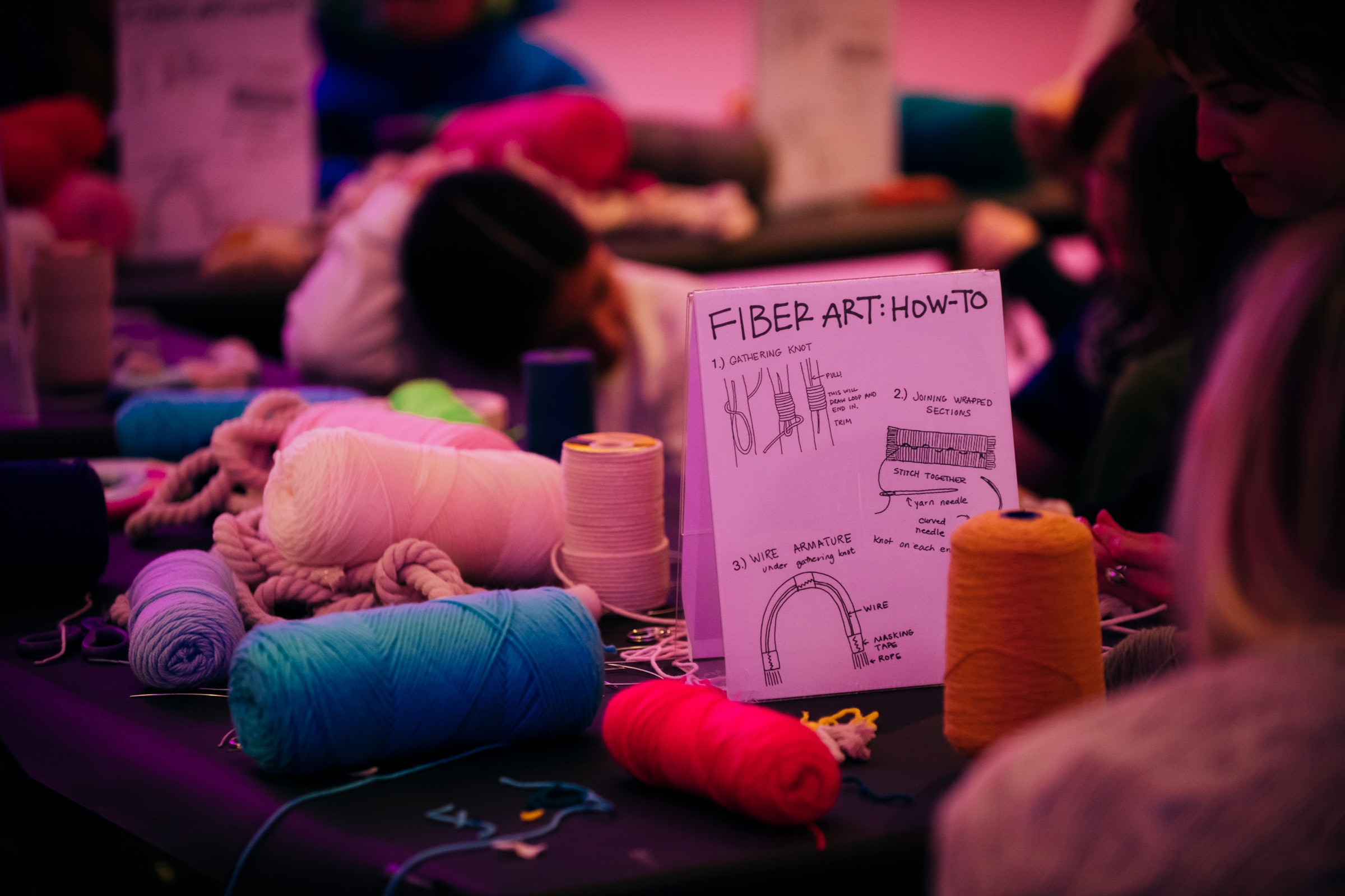 A table with yarn, string, and a Fiber Art: How-To guide; people work on fiber crafts in the background.