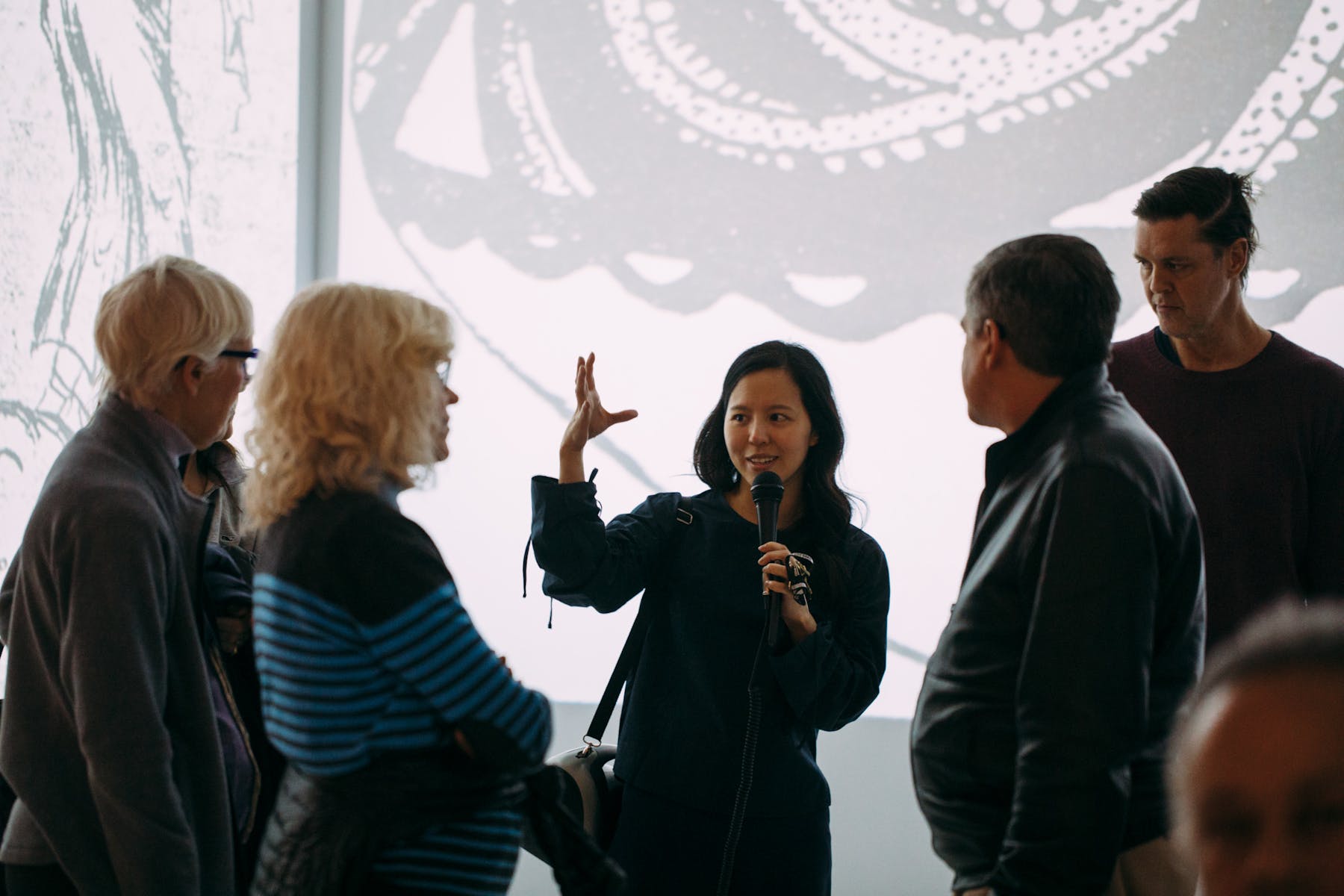 A woman speaks into a microphone that she is hold while raising her other hand and addressing a group of people in a white gallery.