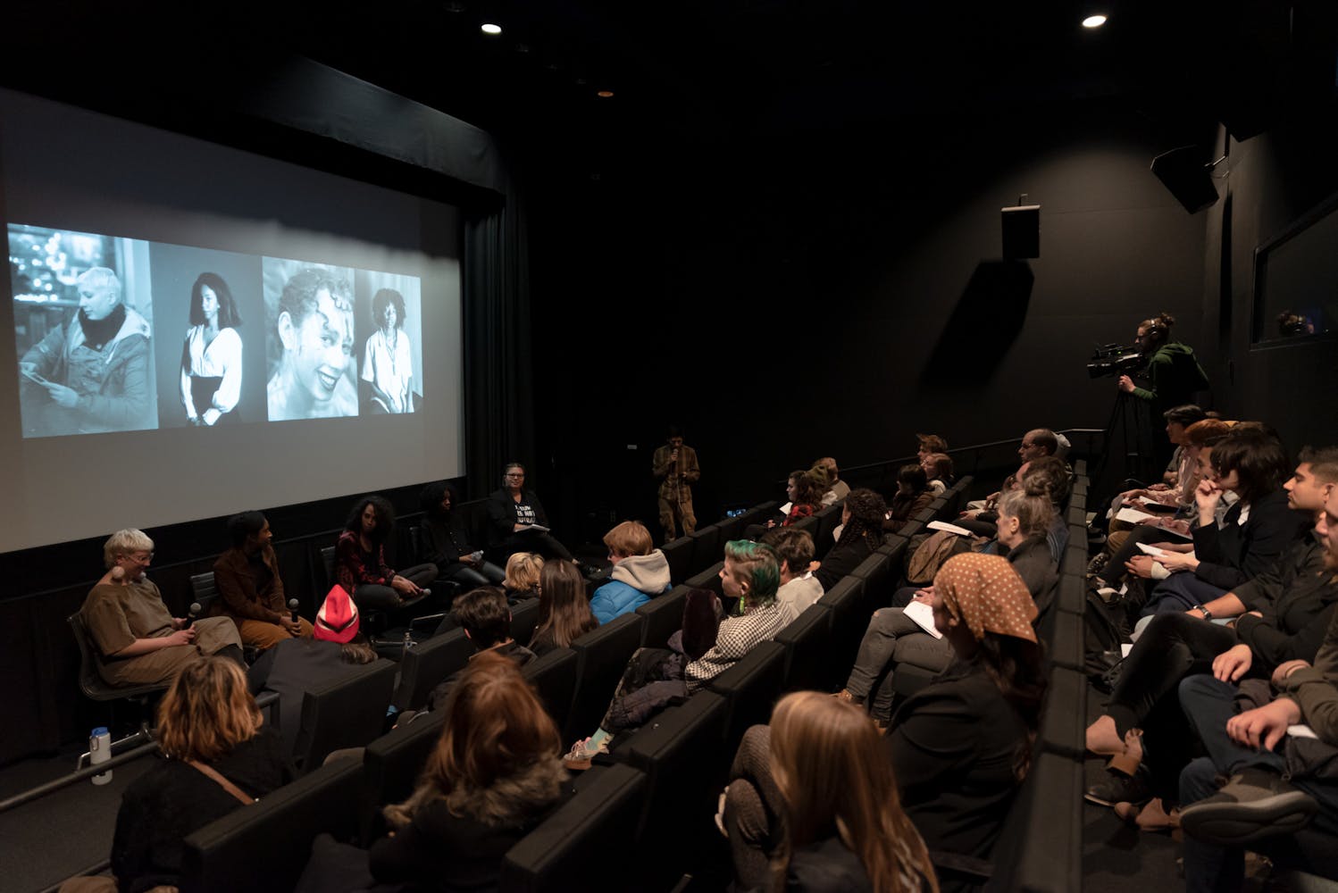 A room of people watch a panel discussion take place in front of a film screen.