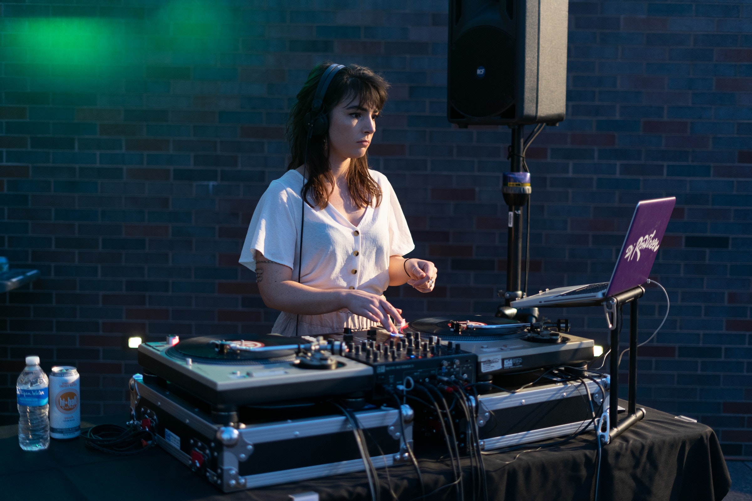 A woman DJ mixes music on turntables outdoors, wearing headphones, with drinks nearby.