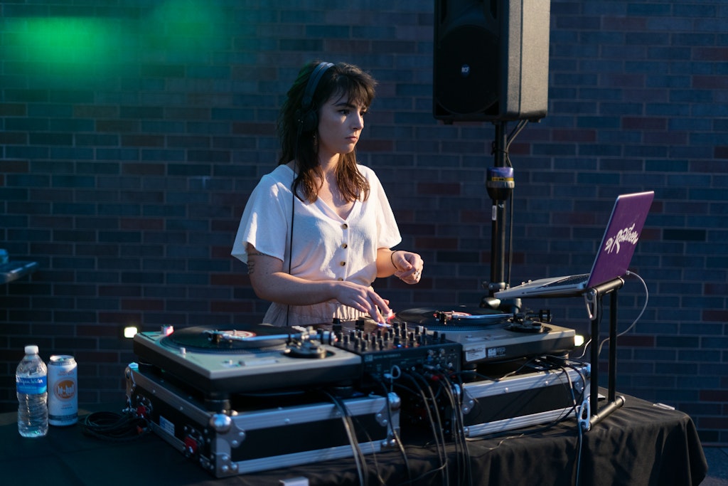 A woman DJ mixes music on turntables outdoors, wearing headphones, with drinks nearby.