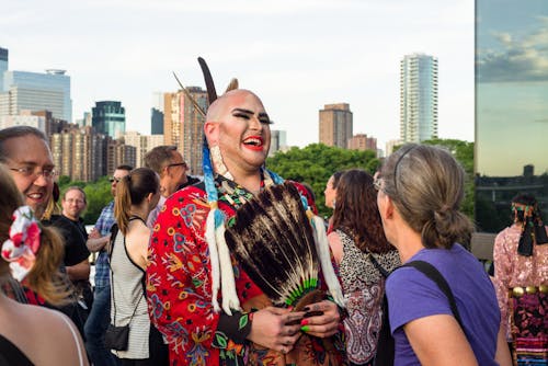 An adult with make-up and Indigenous clothing laughs while talking to another adult on a rooftop terrace.