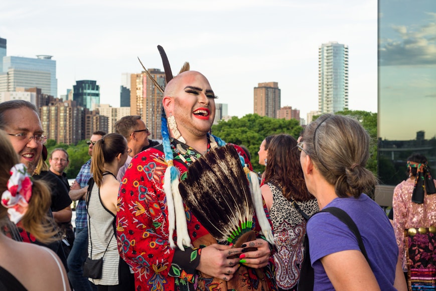 An adult with make-up and Indigenous clothing laughs while talking to another adult on a rooftop terrace.