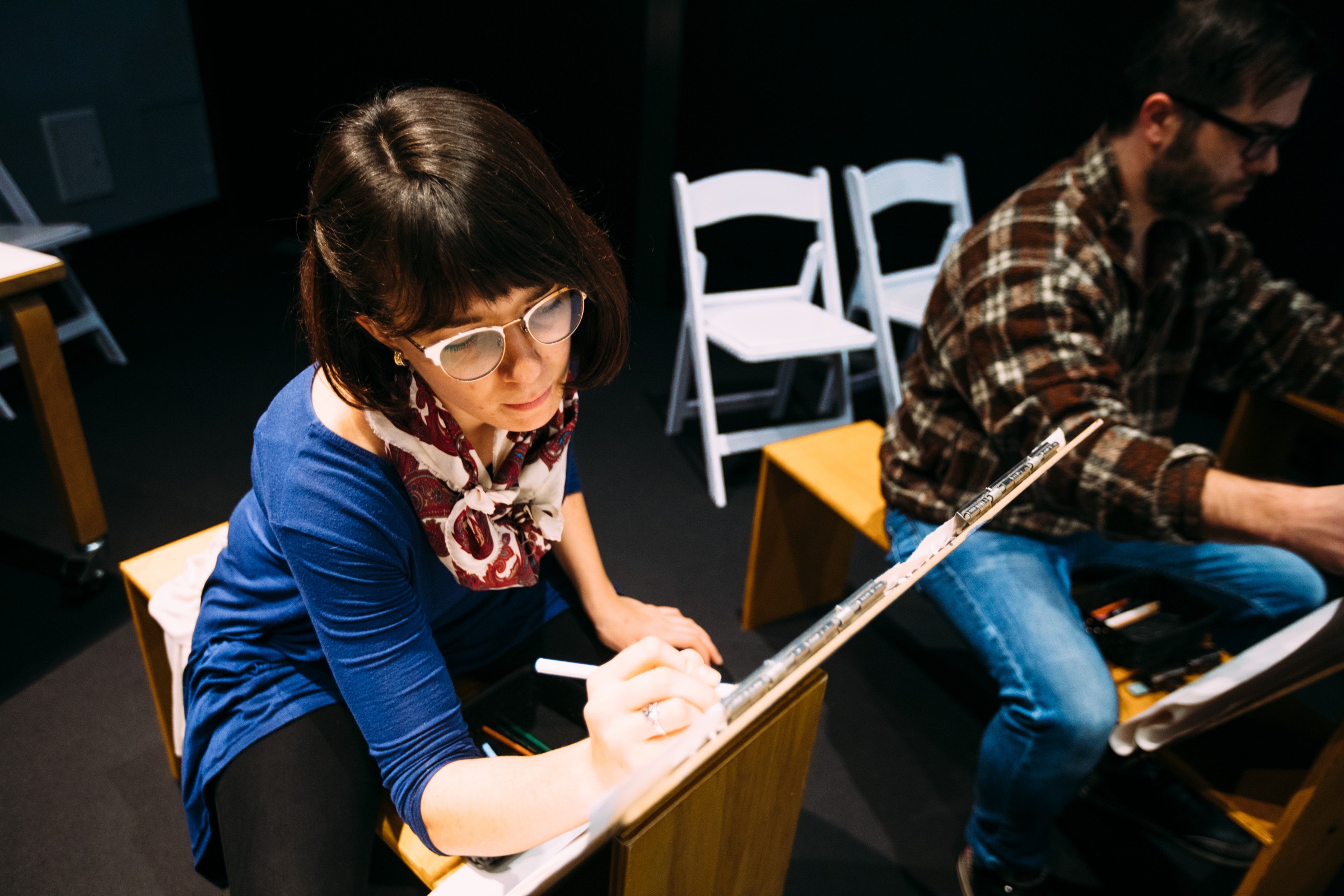 Woman in glasses drawing at an easel, seated next to a man, with empty chairs in the background.