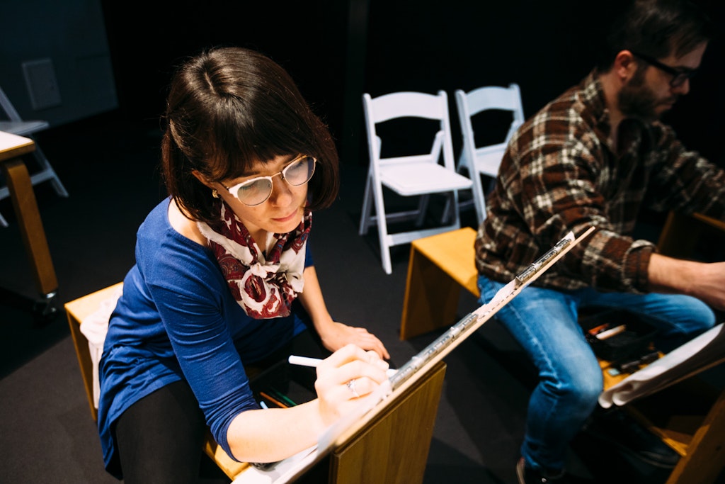 Woman in glasses drawing at an easel, seated next to a man, with empty chairs in the background.