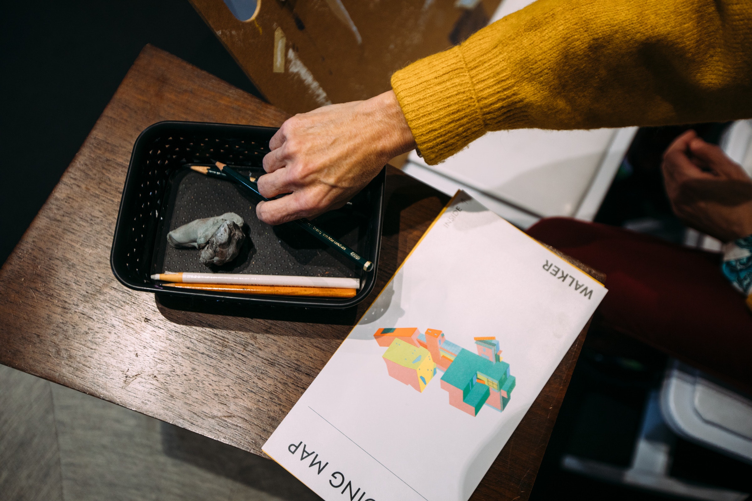 A hand reaches for pencils in a tray next to a colorful folded map of The Walker on a wooden table.