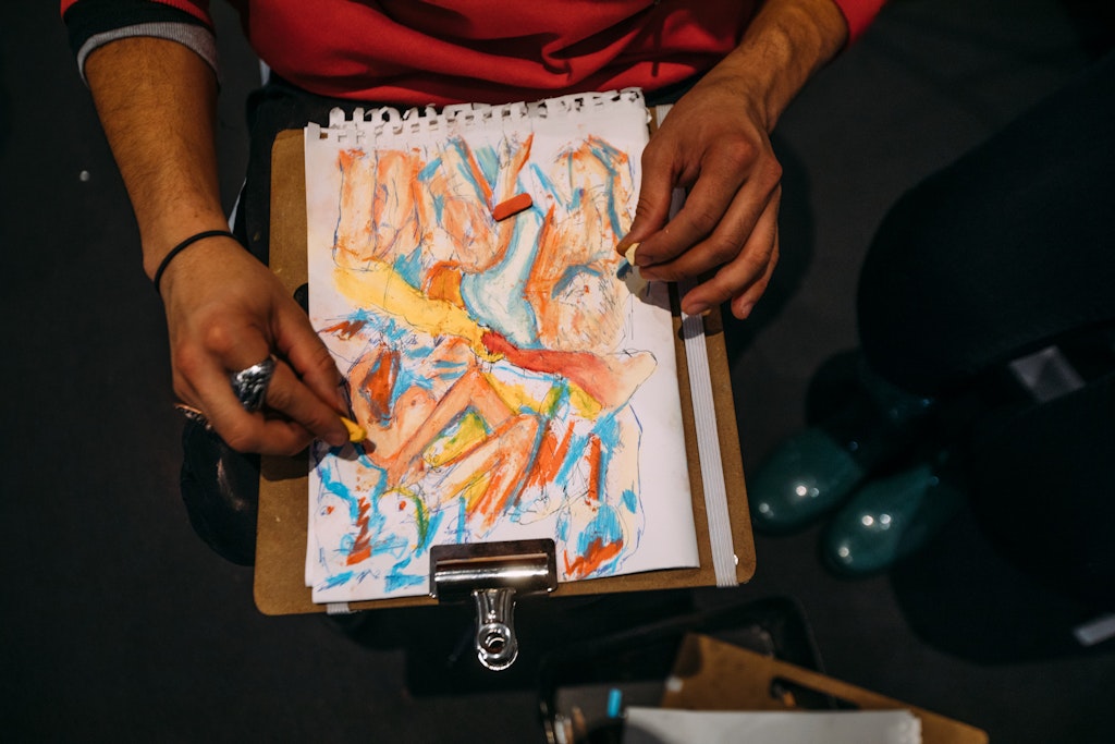 Person drawing a colorful abstract artwork on paper with pastels, holding a clipboard on their lap.