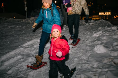 A woman holds the hand of a girl as they snowshoe together at night.