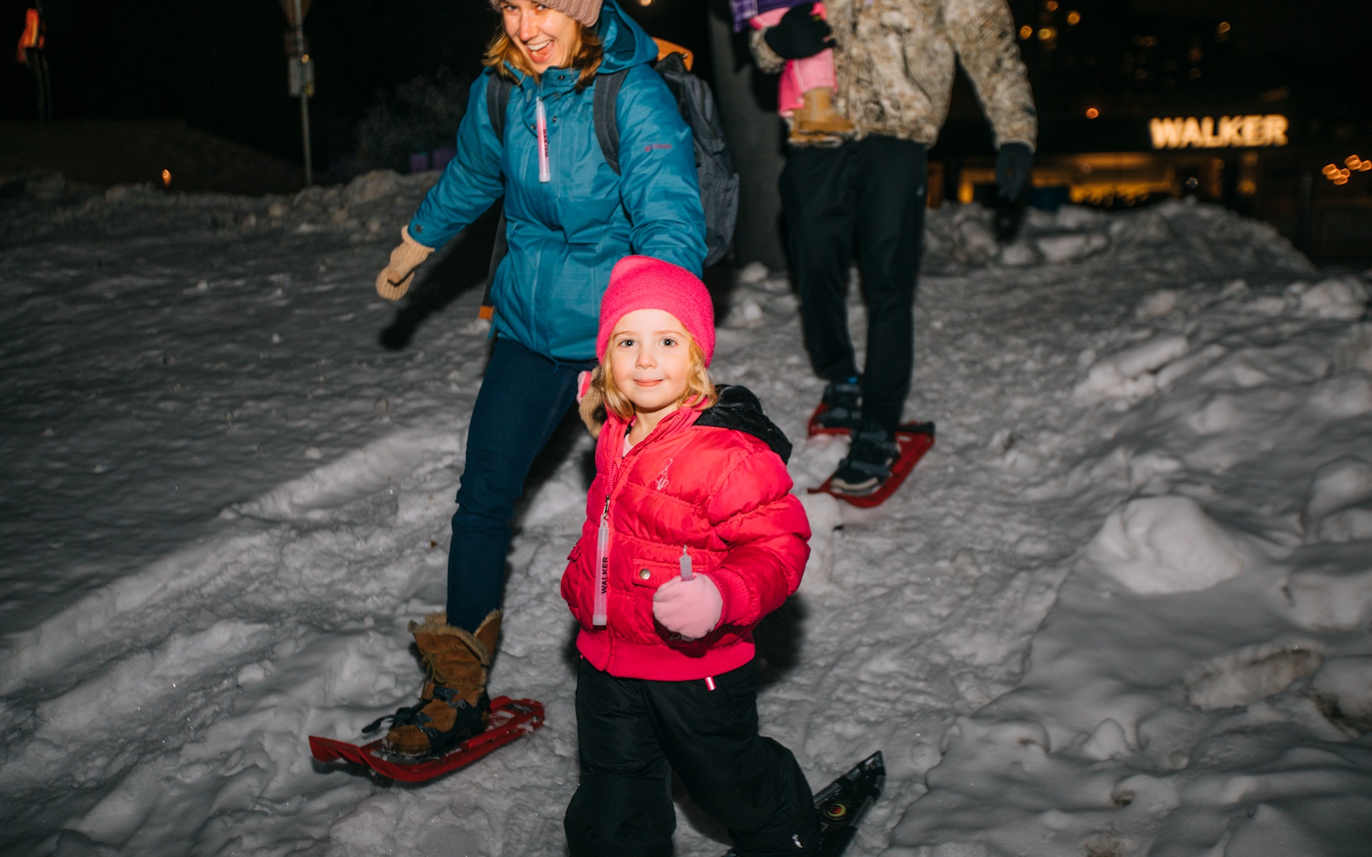 A woman holds the hand of a girl as they snowshoe together at night.