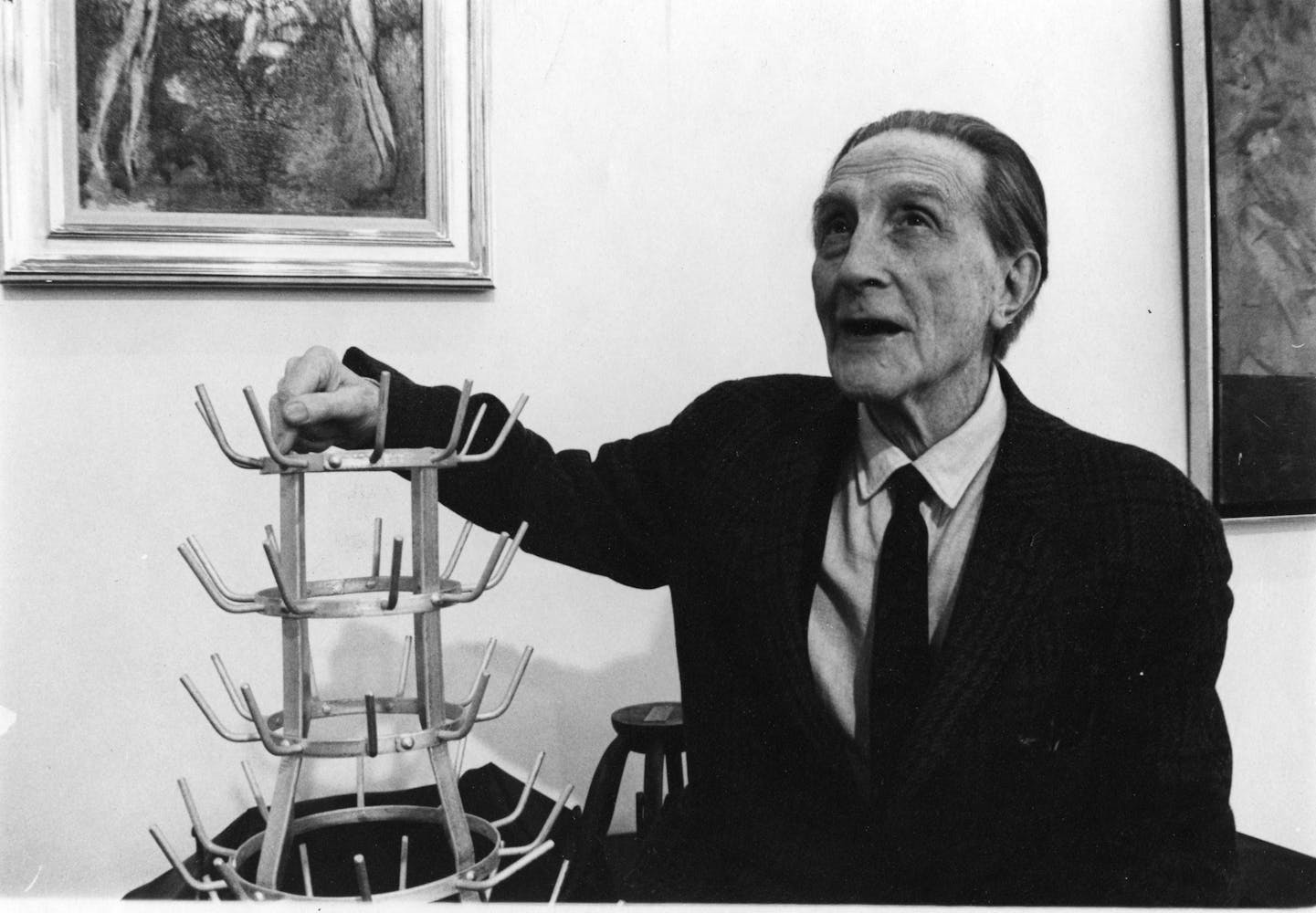An older adult in a suit talks to someone off camera as they rets them arm on a metal bottle drying rack.