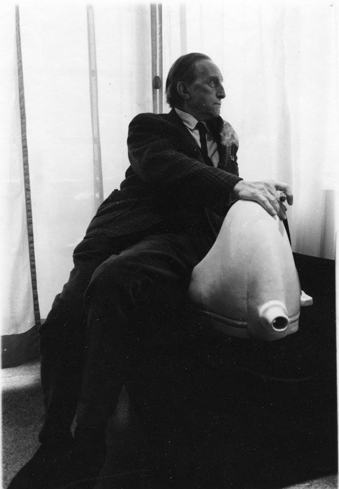 An adult in a suite reclines n a bench next to a cermaic urinal that has been placed on its back.