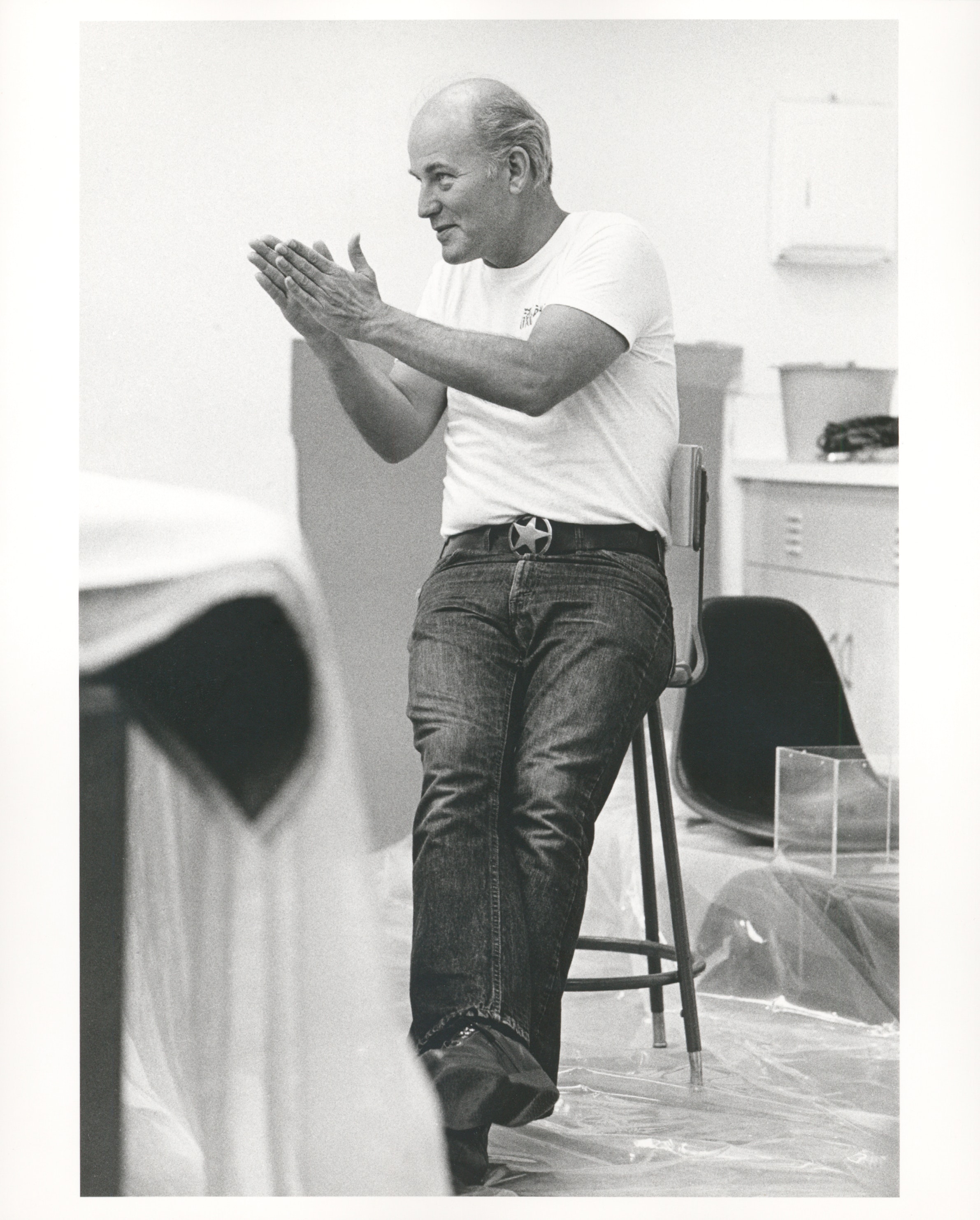 A black and white photograph of a man with light skin and black jeans calpping while sitting on a stool.
