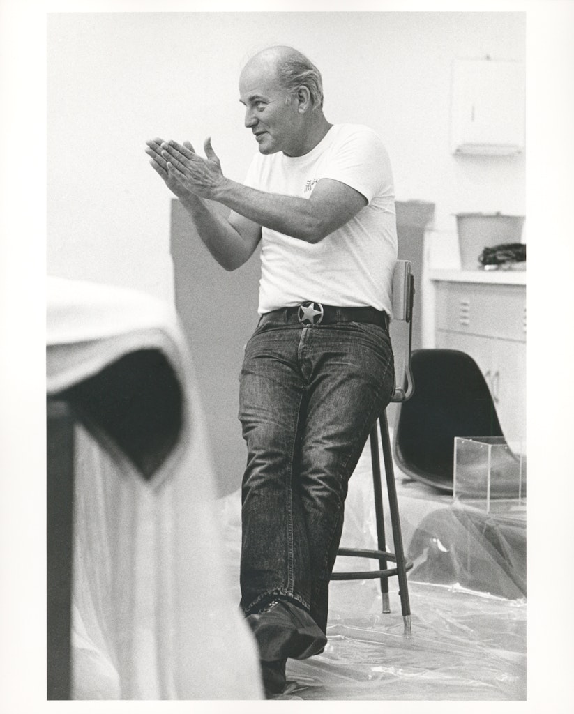 A black and white photograph of a man with light skin and black jeans calpping while sitting on a stool.