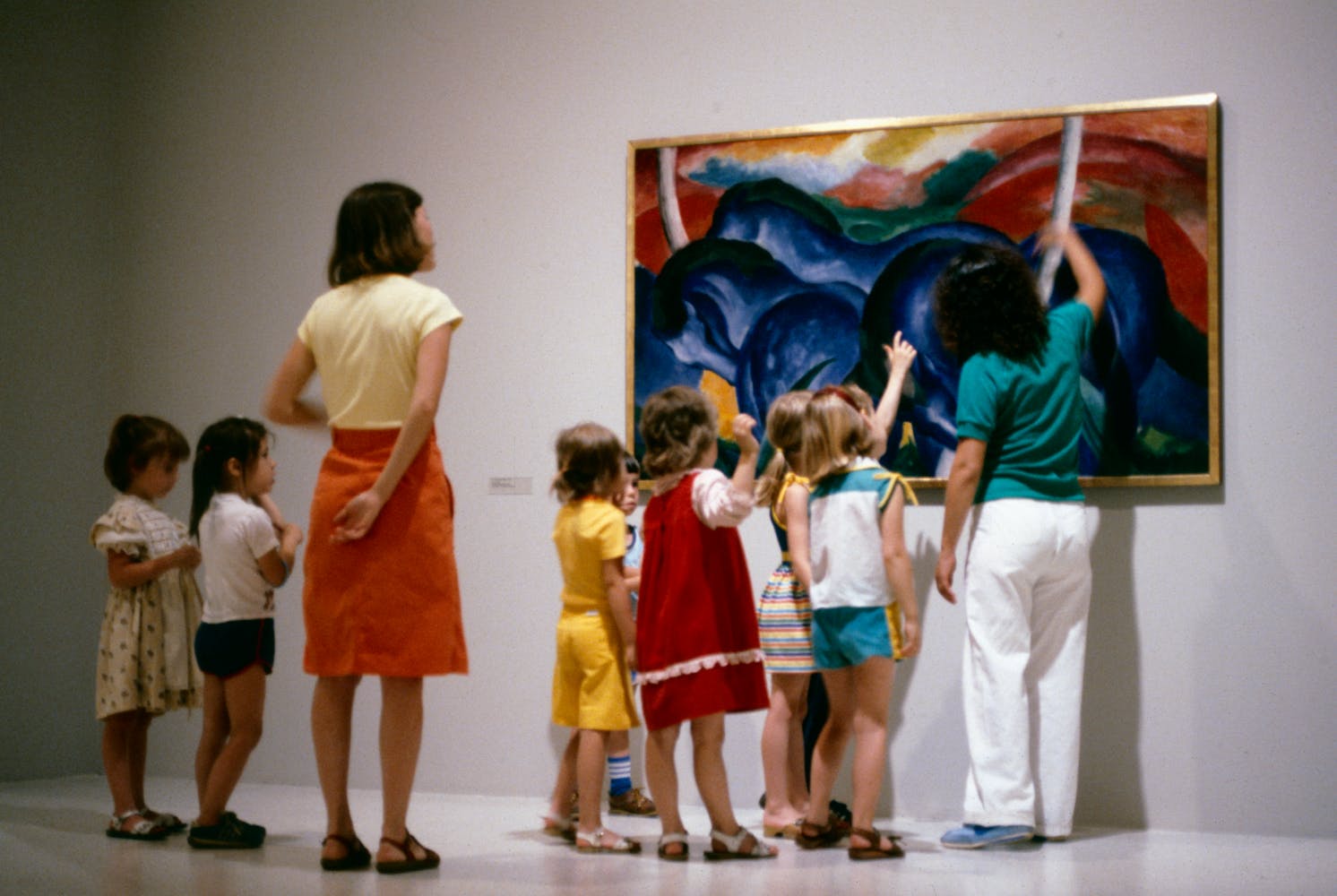 A group of childrend and two adults stand in front of a painting of blue horses.