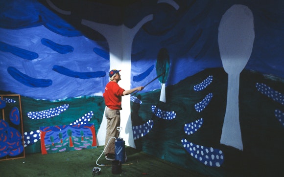 Man in red sweatshirt with a large paint roller working on a mural of colorful trees and sky