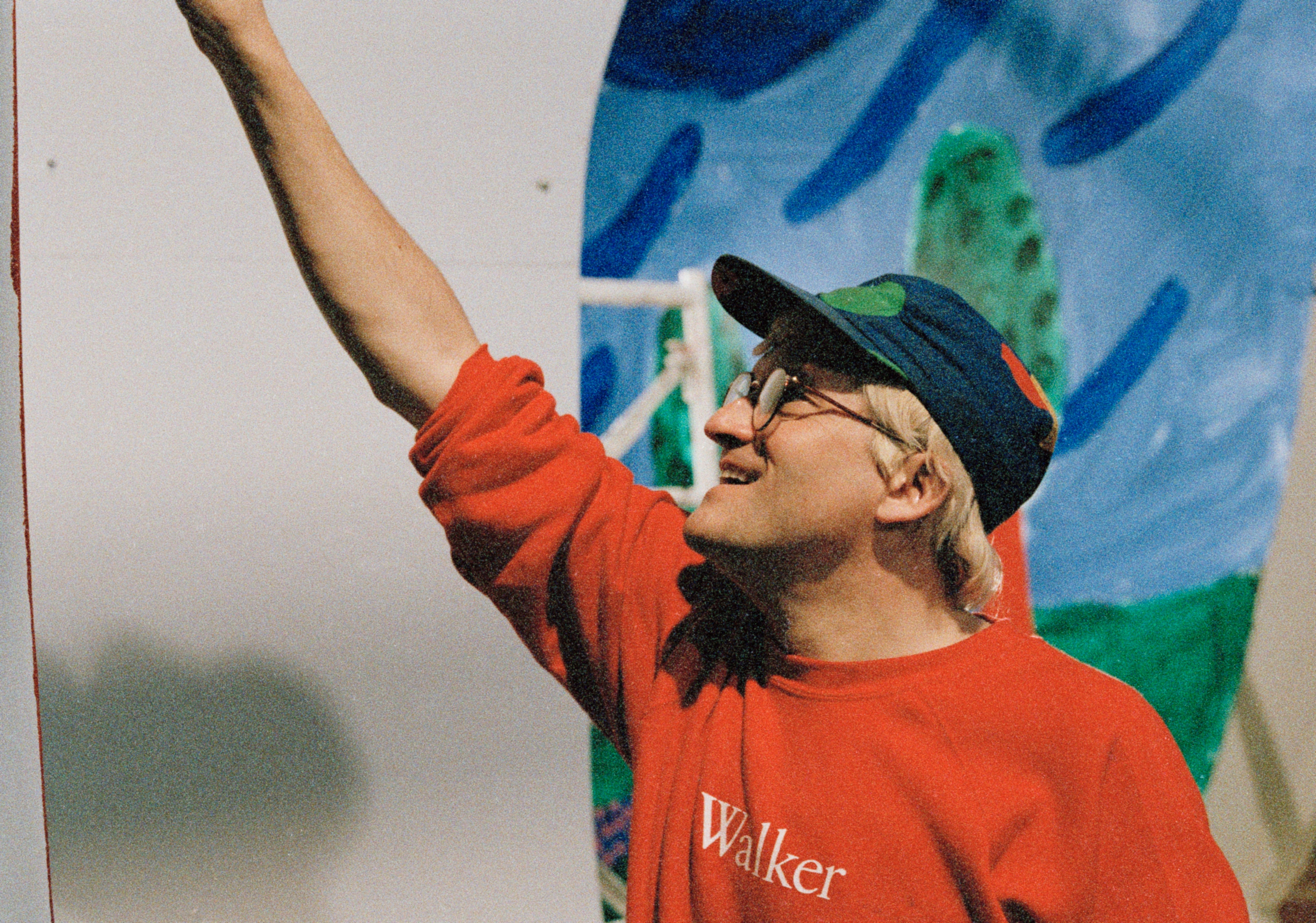 David Hockney, adult with light skin, wearing a red sweatshirt that says 