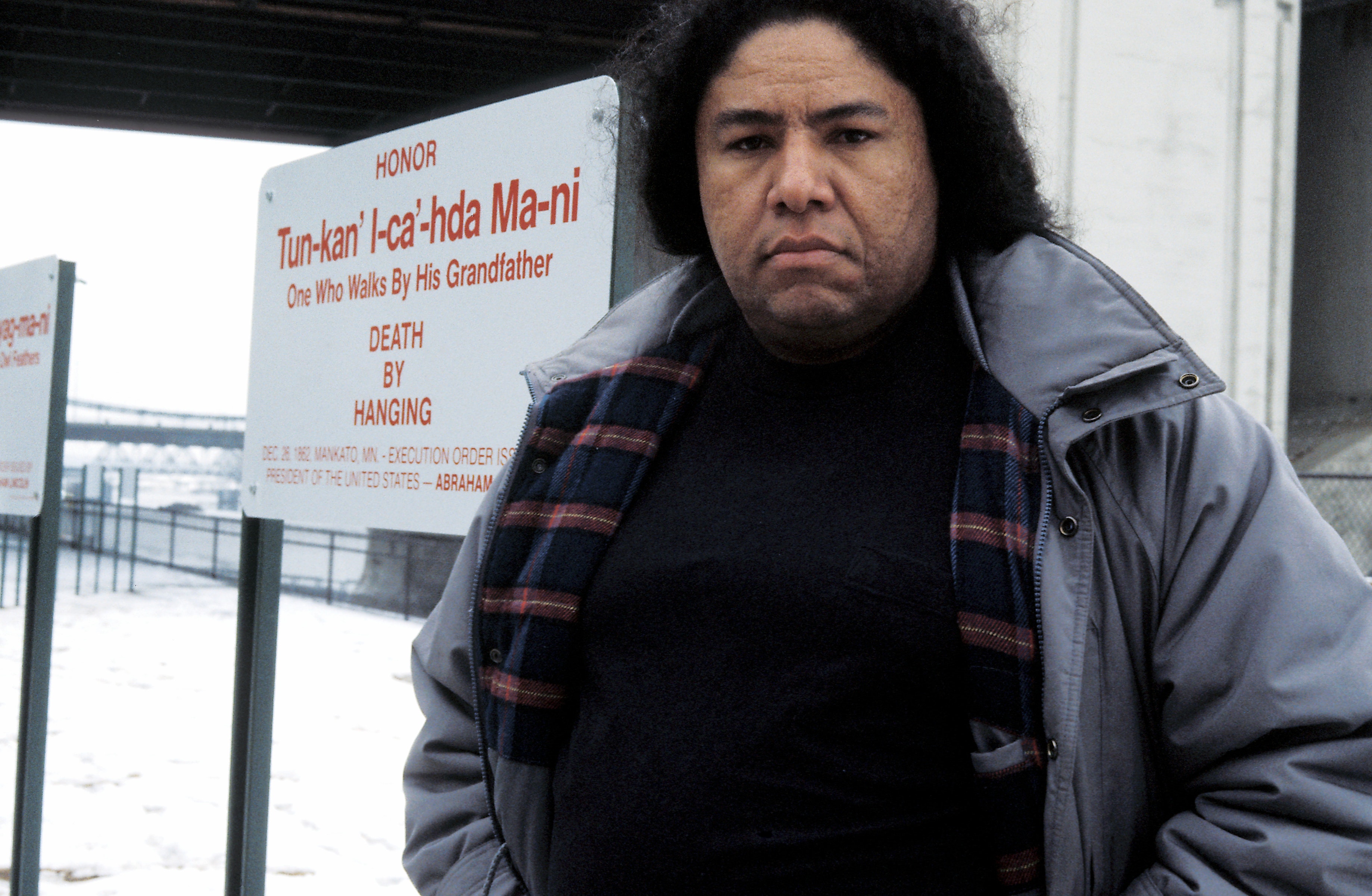 An adult with medium dark skin and black hair, wears a blue winter coat looks at the viewer, standing in front of some signs outside in winter.