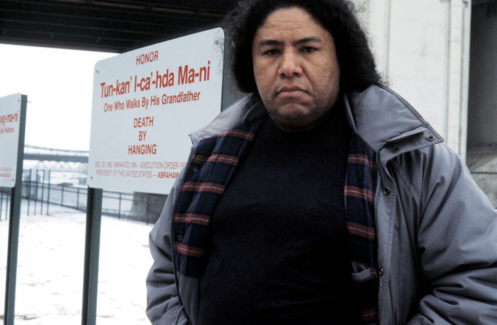 An adult with medium dark skin and black hair, wears a blue winter coat looks at the viewer, standing in front of some signs outside in winter.