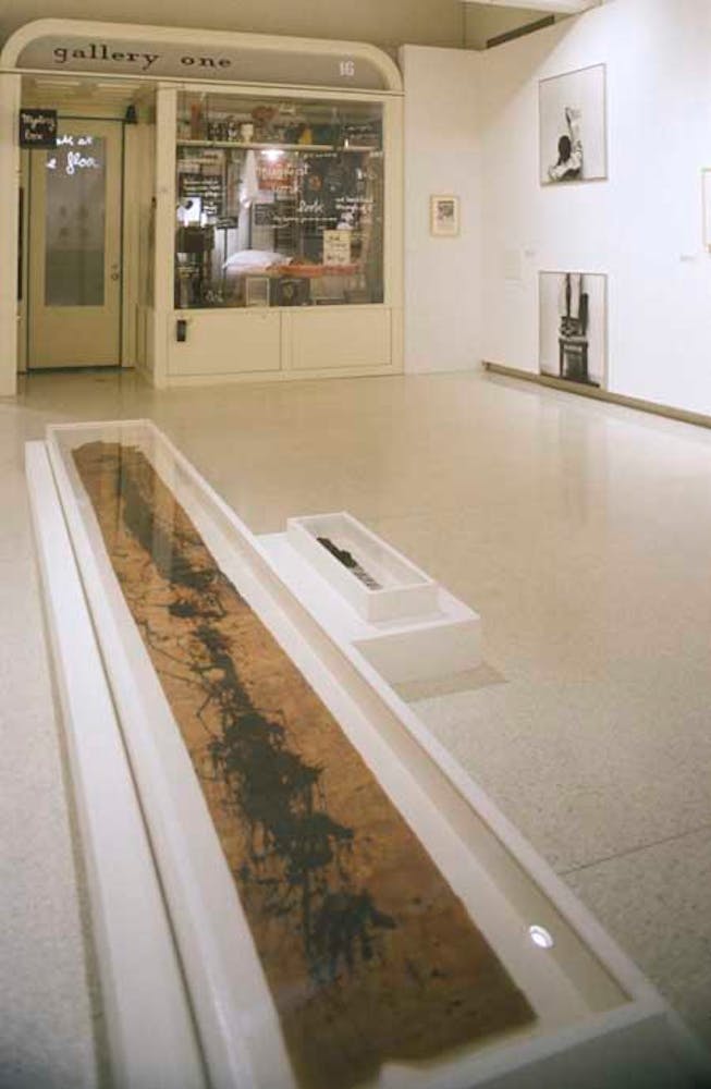 An artwork that is a recreation of the exterior door and window of a shop filled with strange objects is build into the side of a white art gallery.