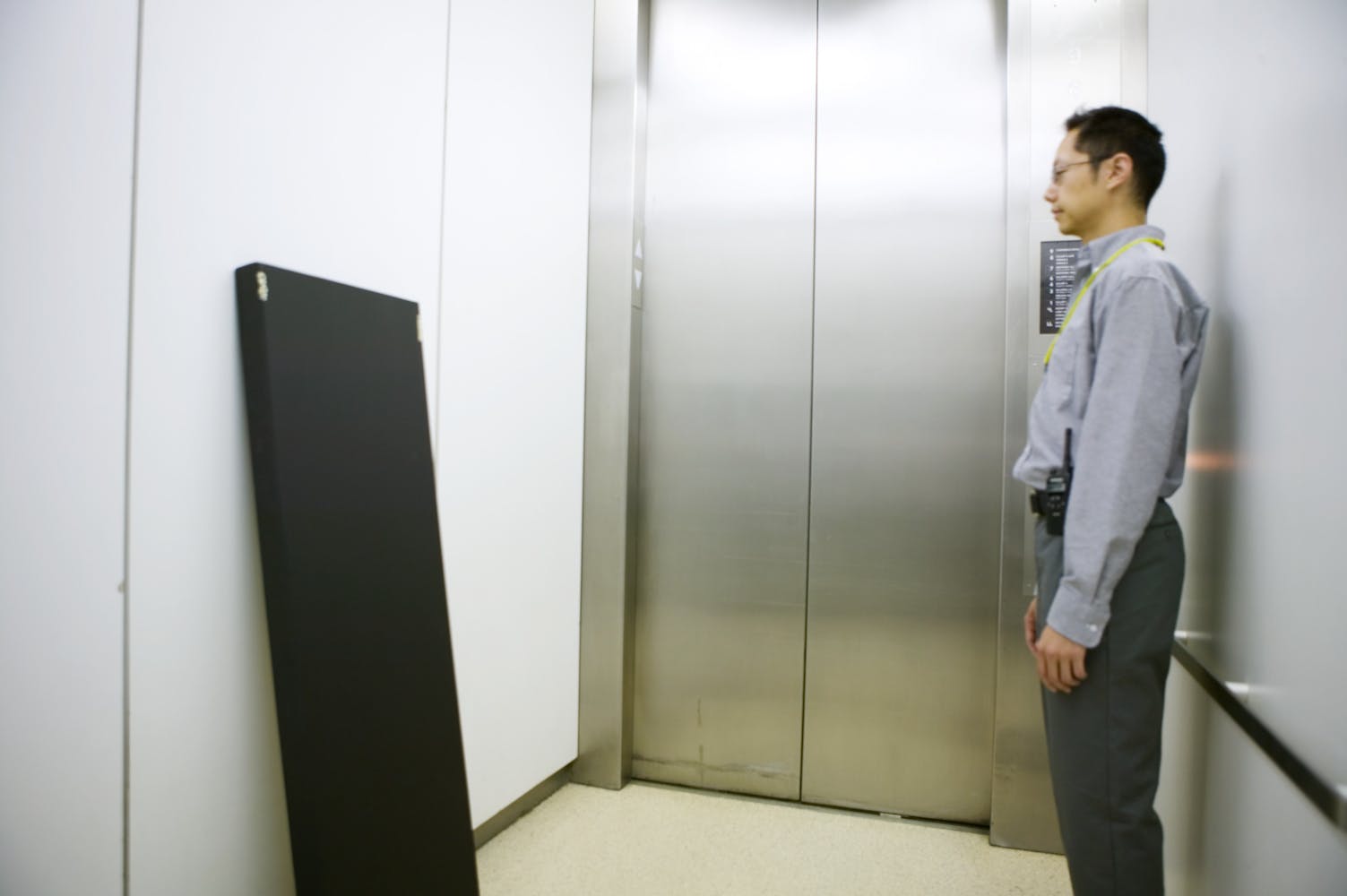 An adult wearing glasses and dress pants and shirt stands in an elevator staring at a black colored canvas leaning against the opposite wall.