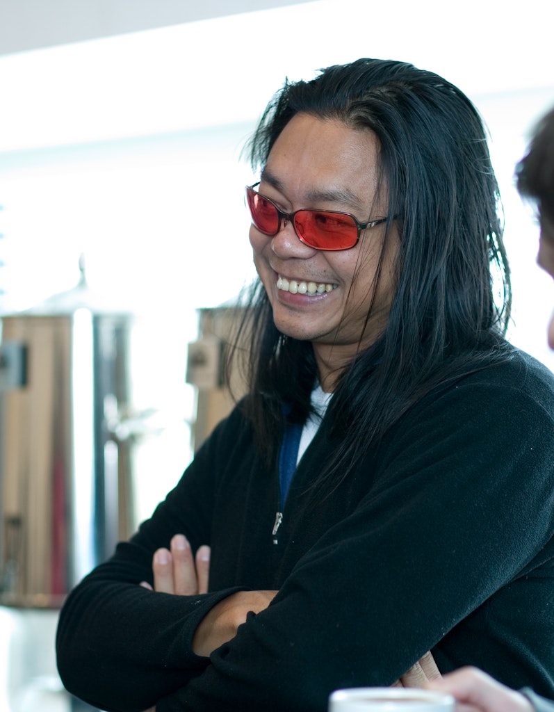 An adult with medium skin and long dark hair, stands crossing their arms and looking towards the right smiling. They are wearing black and red-tinted glasses.
