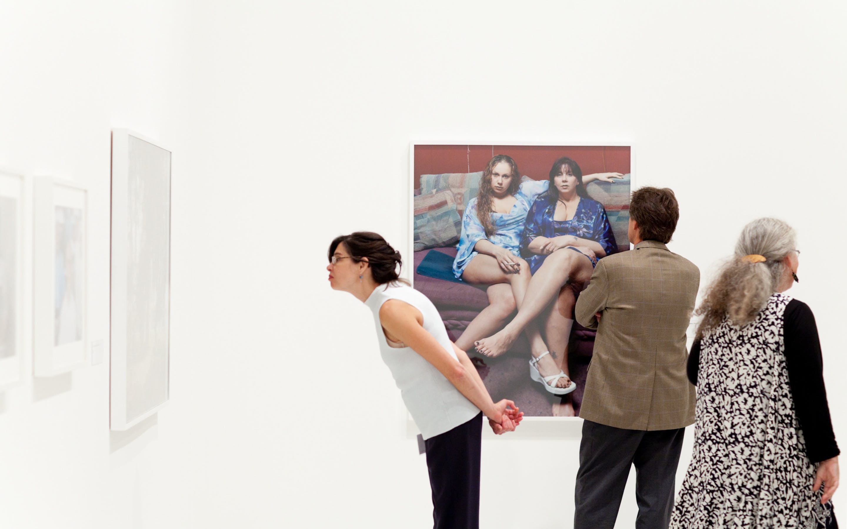 A woman leans forward to look closer at a painting hanging on a gallery wall while two other adults look at another painting on another wall.