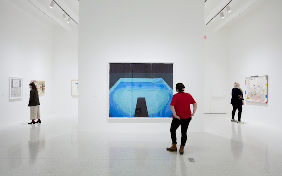 Three people standing in a gallery looking at David Hockneys paintings