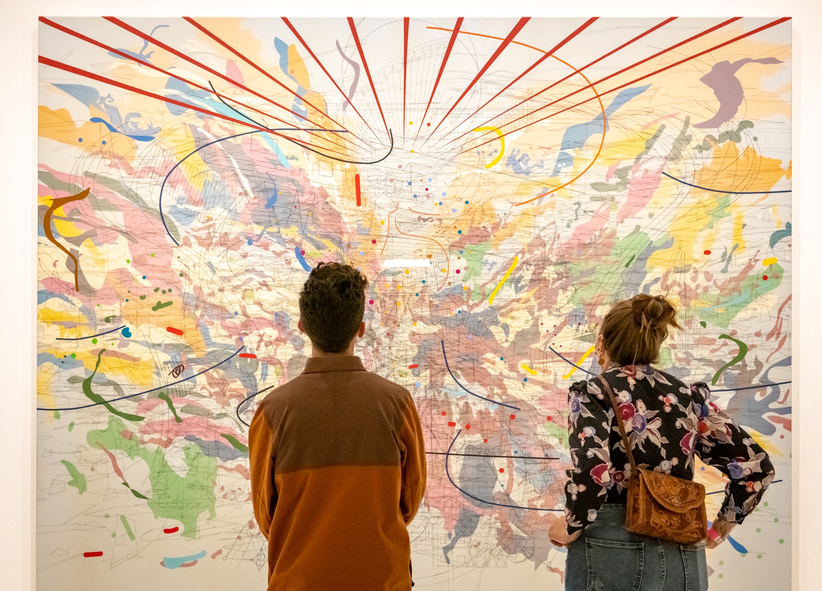 A man and a women stand with their backs to the viewer while examining a painting by Julie Mehretu