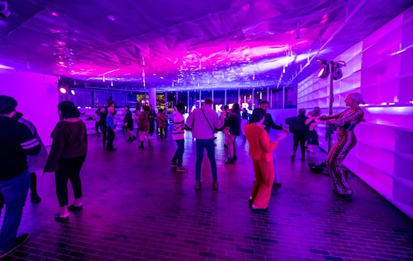 A group of people dance in a museum event space while bathed in purple light.
