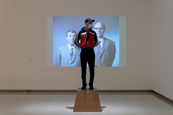Gallery view with man standing on brown wooden platform with projection in the background