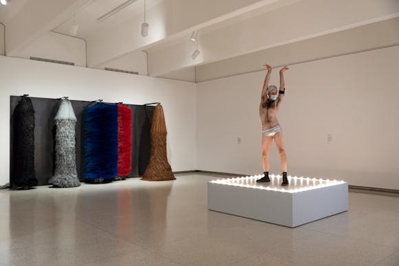 Gallery view with large car wash spinning brushes on wall with man wearing only speedo and shoes dancing on lit platform in foreground